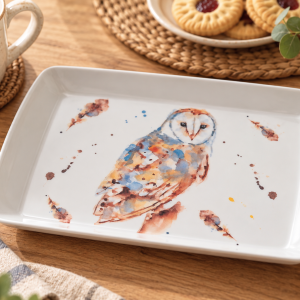 White rectangular plate with a watercolor owl painting, set on a wooden table beside a mug of tea and cookies.