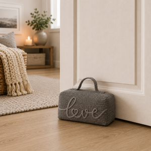 Gray textured bag with the word 'love' in cursive rests on a wooden floor beside a white cabinet in a cozy living space.