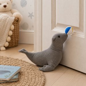 Gray plush seal toy nudges a blue and yellow ball toward a cabinet in a cozy nursery.