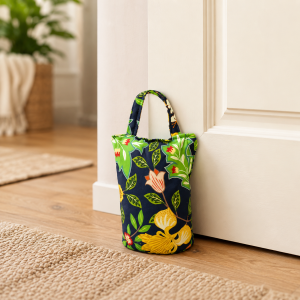 Small fabric tote bag with a bold green, navy, and yellow floral pattern resting against a white cabinet in a bright room on a wooden floor.